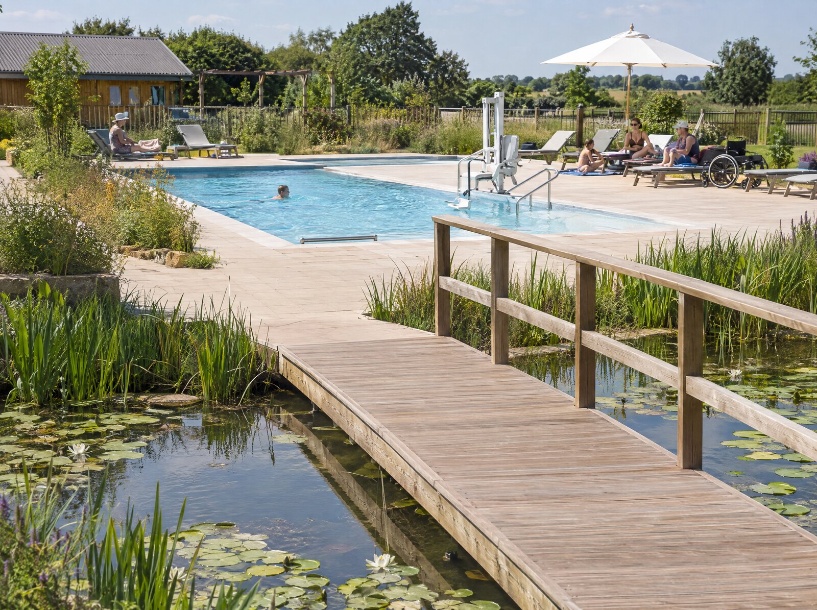 Naturteich mit Schilf und Seerosen und einem breiten Holzsteg, dahinter ein Freibecken mit gepflastertem Sonnendeck, Liegen, Sonnenschirm und einem Poollift.